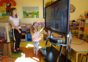 Dziewczynka stoi pod ekranem mobilnym, wskazuje narzędzie pracy, które potrzebuje stomatolog.